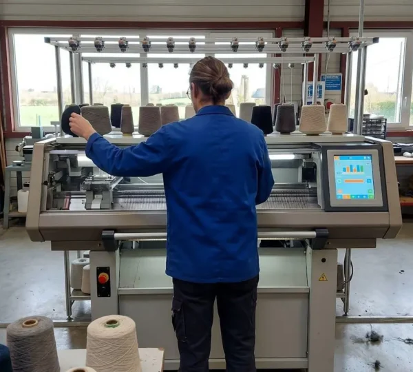Artisan de dos travaillant sur une machine à tricoter industrielle dans un atelier textile français contemporain éclairé par la lumière naturelle