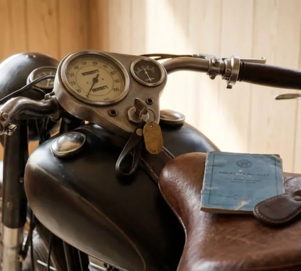 Compteur et selle d'une moto ancienne avec un carnet d'assurance posé dessus, garage baigné de lumière.