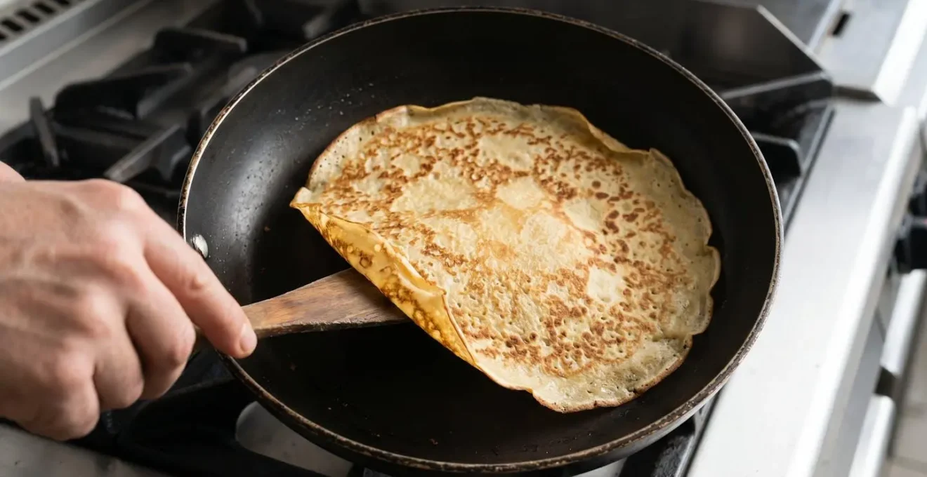 Vue plongeante sur poêle noire avec crêpe dorée dont les bords se décollent, main tenant spatule bois glissée sous le bord avant retournement