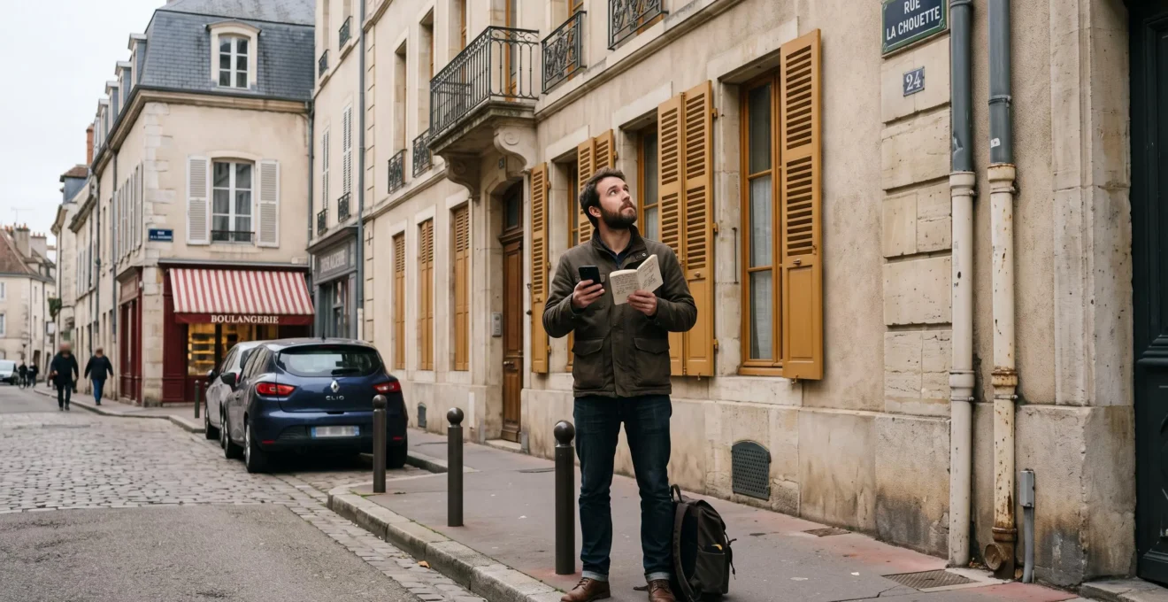 Un homme observe la façade d'un immeuble ancien en pierre dans une rue résidentielle de Dijon, carnet et smartphone en main.